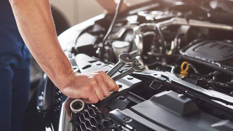 Hand in coveralls holding a wrench over an open engine bay