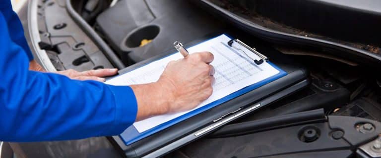 Blue-sleeved hand writing on a clipboard beside an open hood