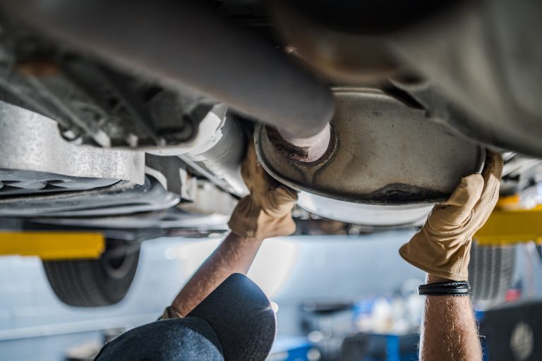 Gloved hands inspecting an exhaust pipe under a lifted vehicle