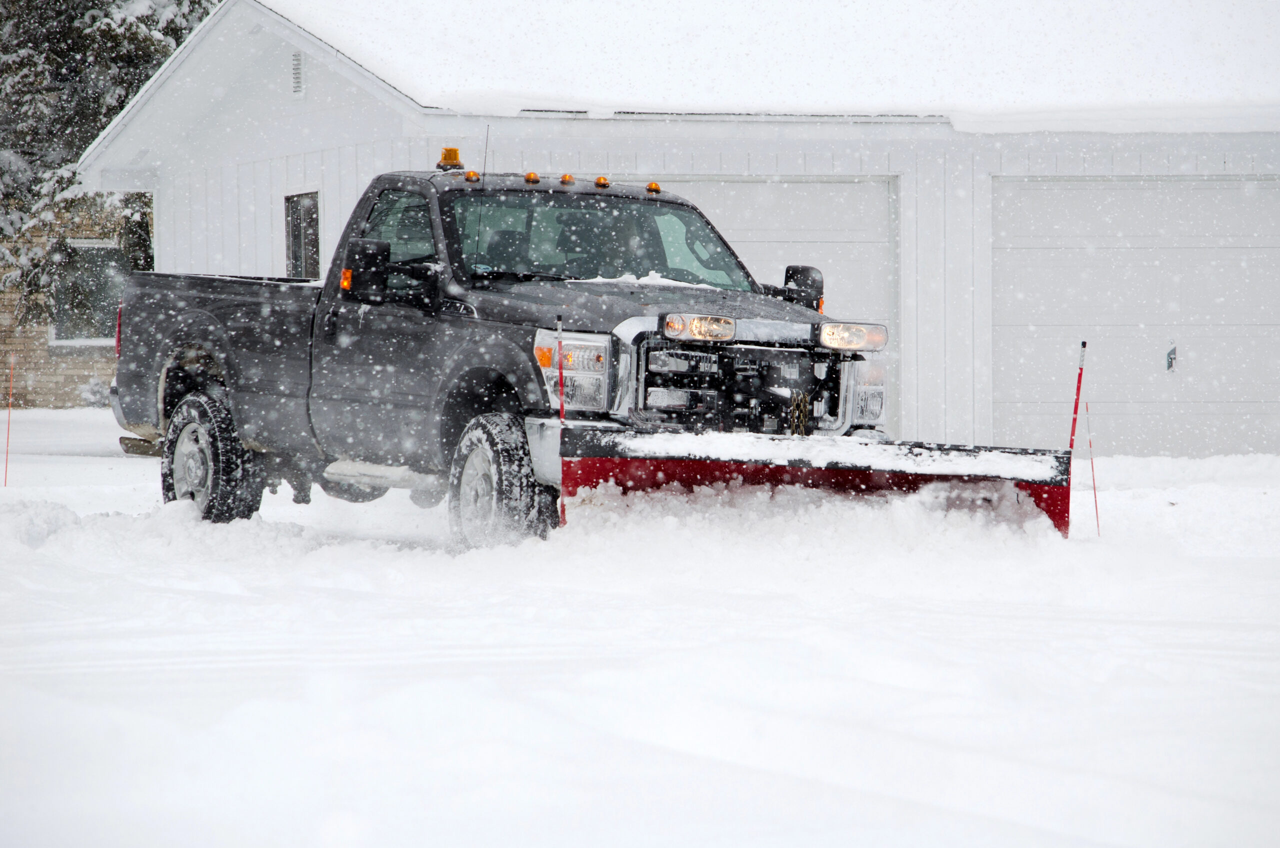 Black pickup truck with red snow plow clearing a driveway during heavy snowfall