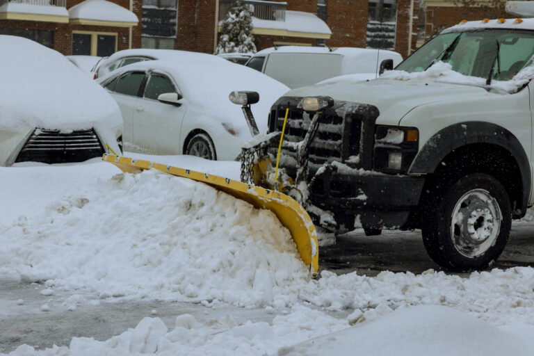 Truck with yellow plow blade clearing deep snow from a parking lot after a storm