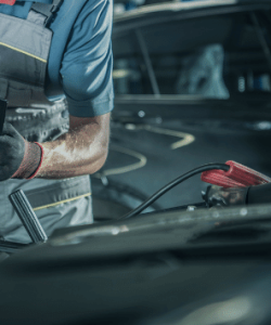 Mechanic holding a clipboard while connecting a jump-start clamp under an open hood