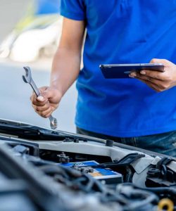 Man in a blue polo holding a wrench and a tablet at an open hood