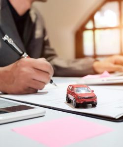 Hand writing on a clipboard beside a model car, laptop and phone on a desk