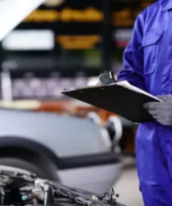 Worker in blue coveralls writing on a clipboard beside a car with the hood up